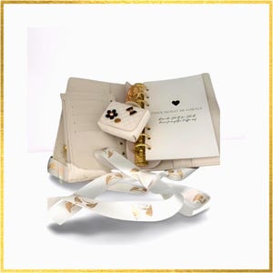 Könnte beinhalten: Cremefarbener Planer mit goldenen Akzenten, geöffnet, um eine Seite mit Text und einem Herzsymbol zu zeigen. Eine kleine, weiße, gesteppte Geldbörse mit Blume und Schleife ist befestigt. Ein weißes Band mit goldenem Muster ist davor drapiert.