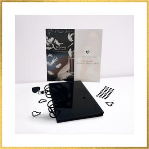 Könnte beinhalten: Ein schwarzes, spiralgebundenes Notizbuch mit glänzendem Einband, zusammen mit zwei Karten und herzförmigen Dekorationen. Die Karten zeigen Text und Illustrationen von Tauben und einem Herzen. Das Notizbuch und die Karten liegen auf weißem Grund.