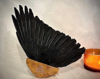 Puede incluir: Un abanico de plumas negras largas fijadas en una base de madera clara pulida. La base de madera presenta un pequeño símbolo de triskelion de triple espiral en color morado grabado en su superficie frontal.