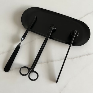 以下が含まれることがあります： 黒い楕円形のトレイに置かれた黒いキャンドルケアセット。セットには、芯ディッパー、芯トリマー、スナッファーが含まれています。ツールは金属製で、キャンドルのメンテナンス用に設計されています。トレイの長さは約25cmです。