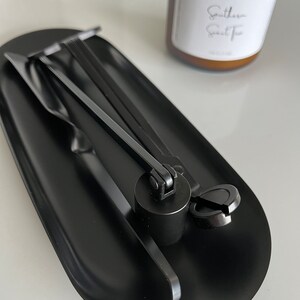 以下が含まれることがあります： 黒いキャンドルアクセサリーセット。芯トリマー、スナッファー、ディッパーが黒い楕円形のトレイに置かれています。背景には、白いラベルが付いた茶色のキャンドルがあります。ラベルには「Southern Sweet Tea」と書かれています。