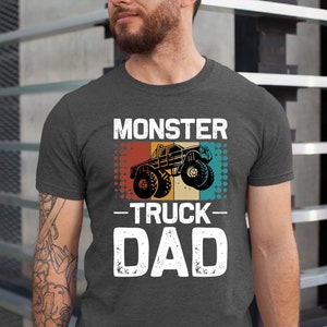 May include: A dark gray t-shirt with a vintage-style graphic of a monster truck. The text "Monster Truck Dad" is printed in white on the shirt.