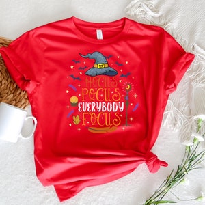 May include: A red t-shirt with a Halloween-themed graphic. The design includes a witch's hat, bats, and the phrase "Hocus Pocus Everybody Focus" in colorful lettering. A white mug and decorative items are in the background.