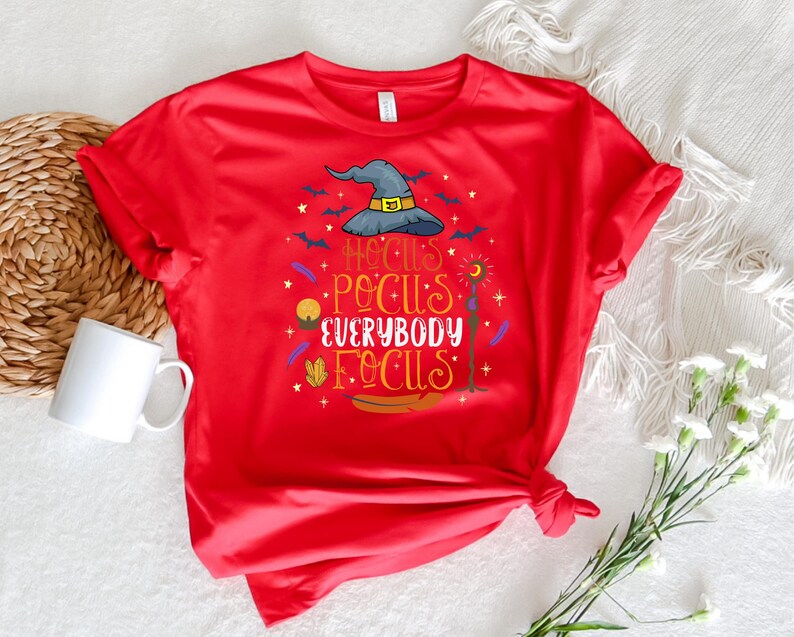 May include: A red t-shirt with a Halloween-themed graphic. The design includes a witch's hat, bats, and the phrase "Hocus Pocus Everybody Focus" in colorful lettering. A white mug and decorative items are in the background.