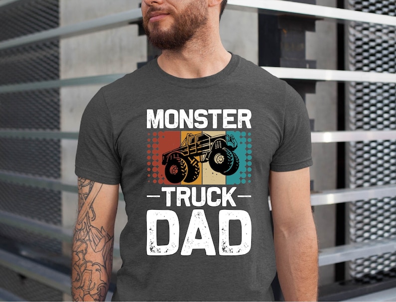 May include: A dark gray t-shirt with a vintage-style graphic of a monster truck. The text "Monster Truck Dad" is printed in white on the shirt.