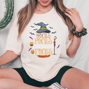 May include: Cream-colored t-shirt with the phrase "Hocus Pocus Everybody Focus" in orange and purple. The design includes a witch's hat, bats, feathers, and other Halloween-themed graphics. The shirt is a casual, oversized fit.