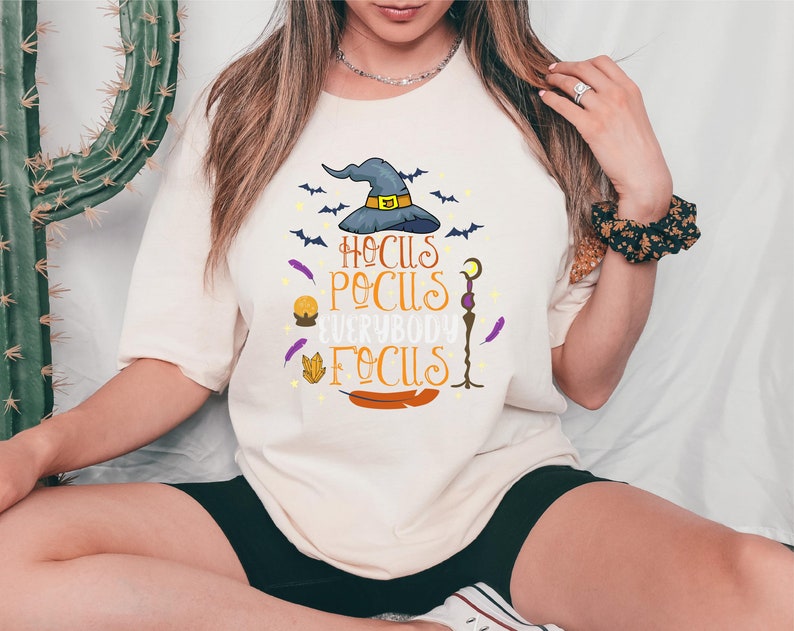 May include: Cream-colored t-shirt with the phrase "Hocus Pocus Everybody Focus" in orange and purple. The design includes a witch's hat, bats, feathers, and other Halloween-themed graphics. The shirt is a casual, oversized fit.