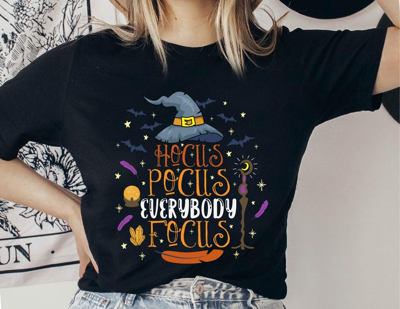 May include: Black t-shirt with a Halloween-themed graphic. The design features a witch's hat, bats, stars, and the text "Hocus Pocus Everybody Focus" in orange and yellow. Additional elements include a crystal ball, feathers, and a candle.