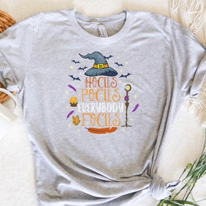 May include: A light gray t-shirt featuring a colorful graphic with a witch hat, bats, and the text "Hocus Pocus Everybody Focus". The design includes a crystal, feather, and a staff. A fun and festive design for Halloween.