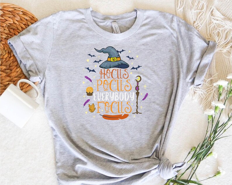 May include: A light gray t-shirt featuring a colorful graphic with a witch hat, bats, and the text "Hocus Pocus Everybody Focus". The design includes a crystal, feather, and a staff. A fun and festive design for Halloween.