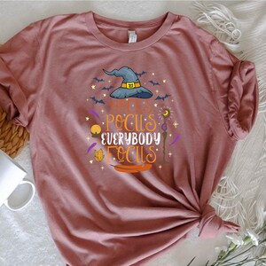 May include: A dusty rose t-shirt with a Halloween-themed graphic. The design features a witch's hat, bats, and the phrase "Hocus Pocus Everybody Focus" in orange and purple lettering. The shirt is knotted at the bottom.