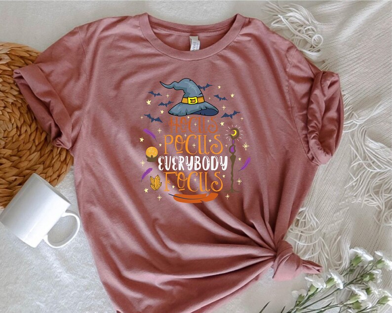 May include: A dusty rose t-shirt with a Halloween-themed graphic. The design features a witch's hat, bats, and the phrase "Hocus Pocus Everybody Focus" in orange and purple lettering. The shirt is knotted at the bottom.