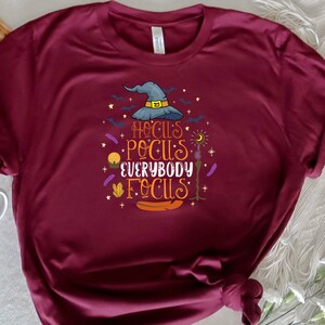 May include: Burgundy t-shirt with a colorful graphic that reads "Hocus Pocus Everybody Focus." The design includes a witch's hat, bats, stars, and other Halloween-themed elements. The shirt is made of a soft material.
