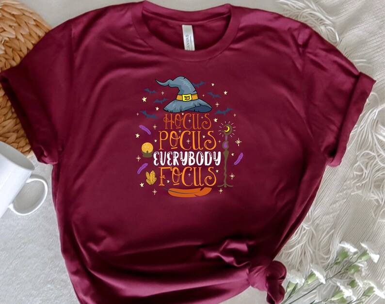 May include: Burgundy t-shirt with a colorful graphic that reads "Hocus Pocus Everybody Focus." The design includes a witch's hat, bats, stars, and other Halloween-themed elements. The shirt is made of a soft material.