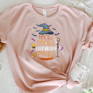 May include: A light pink t-shirt with a Halloween-themed graphic. The design features a witch's hat, bats, and the phrase "Hocus Pocus Everybody Focus" in various colors. The shirt is made of soft material.
