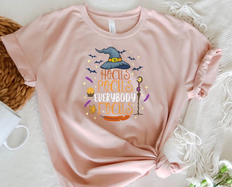 May include: A light pink t-shirt with a Halloween-themed graphic. The design features a witch's hat, bats, and the phrase "Hocus Pocus Everybody Focus" in various colors. The shirt is made of soft material.
