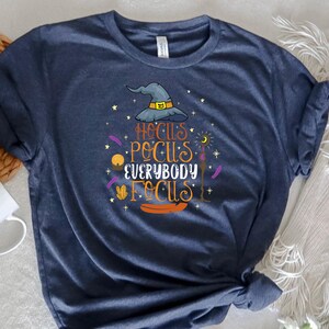 May include: Navy blue t-shirt with a Halloween-themed graphic. The design features a witch's hat, stars, and the text "Hocus Pocus Everybody Focus" in orange and yellow. The shirt is made of soft material.