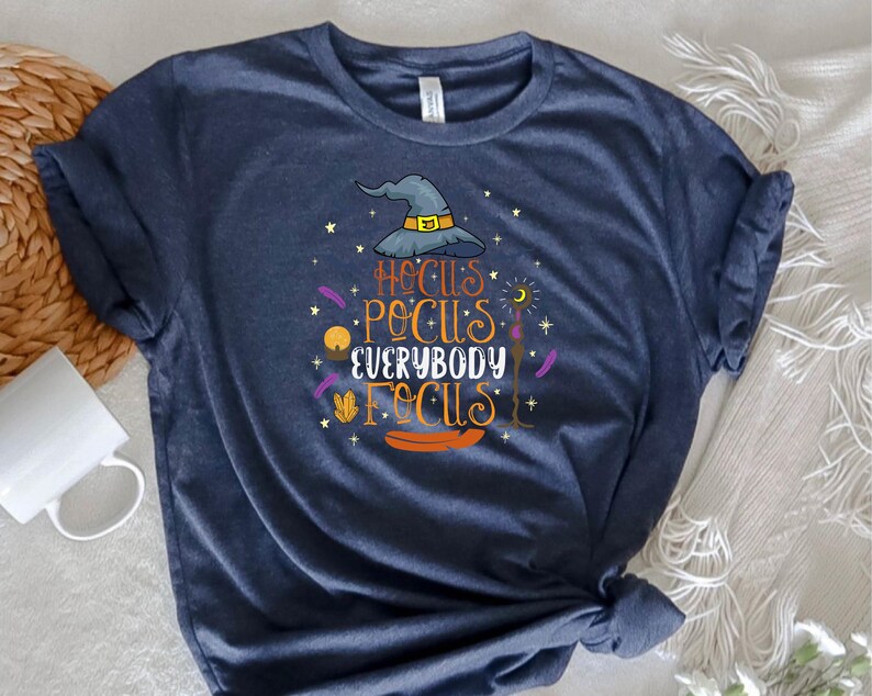 May include: Navy blue t-shirt with a Halloween-themed graphic. The design features a witch's hat, stars, and the text "Hocus Pocus Everybody Focus" in orange and yellow. The shirt is made of soft material.