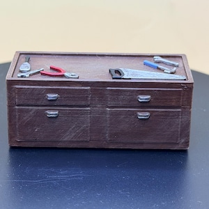 May include: A miniature brown wooden workbench featuring two drawers with silver handles. The top surface holds several small tools, including a red-handled pair of pliers, a hand saw with a black handle, and various metallic wrenches.