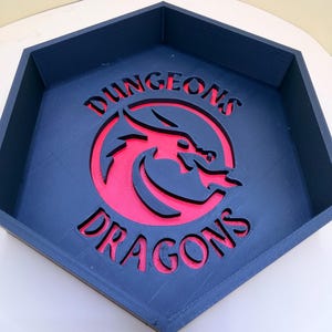 May include: A hexagonal dark blue tray featuring a cutout of a dragon head emblem in the center. The text DUNGEONS is positioned above the dragon, and DRAGONS is below it. The base of the tray is a vibrant red, visible through the cutouts.