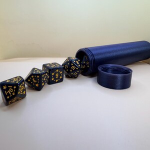 May include: A set of dark blue polyhedral dice featuring gold-colored numbers and small star motifs. The dice are arranged in a line next to a matching dark blue cylindrical storage tube with a separate circular lid placed beside it.