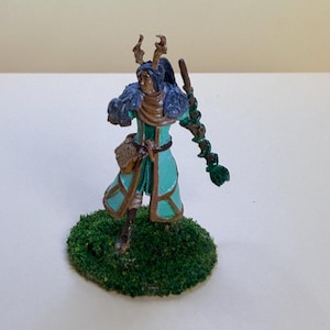 Peut inclure: Une figurine miniature peinte représentant un personnage avec des bois, portant un manteau bleu sarcelle à bordure marron, un col en fourrure bleu foncé et une sacoche marron. Le personnage tient un long bâton entouré de vignes vertes sur un socle.