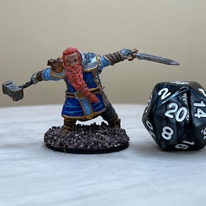 Puede incluir: Una figura miniatura pintada de un guerrero enano sosteniendo un martillo y una espada. El enano lleva una armadura azul y dorada y tiene una larga barba roja. La figura está de pie sobre una base marrón. Un dado negro de 20 caras está en el fondo.