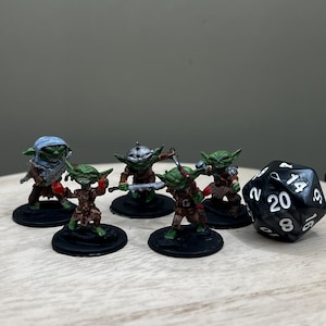 Puede incluir: Cinco miniaturas de goblins verdes y marrones con detalles pintados, de pie sobre bases negras. Un dado negro de 20 caras con números blancos está en primer plano.