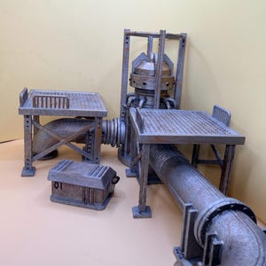 Könnte beinhalten: Ein industrielles Miniatur-Geländeset mit zwei erhöhten Metallplattformen mit Gitteroberflächen, die durch eine zentrale Rohrstruktur mit einem zylindrischen Tank verbunden sind. Eine kleine rechteckige Aufbewahrungskiste mit der Nummer 01 ist enthalten.