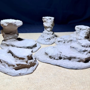 Puede incluir: Un conjunto de cinco piezas de terreno impresas en 3D, de color gris claro y sin pintar, que presentan afloramientos rocosos y formaciones de piedra texturizadas. Estos elementos de paisaje en miniatura muestran líneas de impresión.