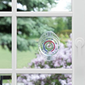May include: Clear, oval window thermometer with red and blue temperature markings. The thermometer is mounted on a white window frame, with a blurred outdoor background of green trees and purple flowers.