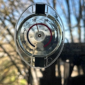 Może przedstawiać: Przezroczysty, owalny termometr okienny z czarnymi akcentami. Termometr wyświetla skale Celsjusza i Fahrenheita, z czerwoną igłą wskazującą aktualną temperaturę. Temperatura wynosi obecnie 15,5 stopnia Celsjusza.