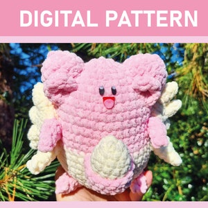 Op de afbeelding: Een roze en crèmekleurig gehaakt amigurumi-speelgoed met een glimlachend gezicht en een roze tong die uitsteekt. Het speelgoed heeft een staart en vleugels.