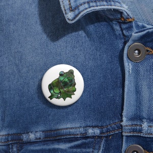 May include: A white button featuring a green and blue frog illustration. The frog is sitting on a white background.