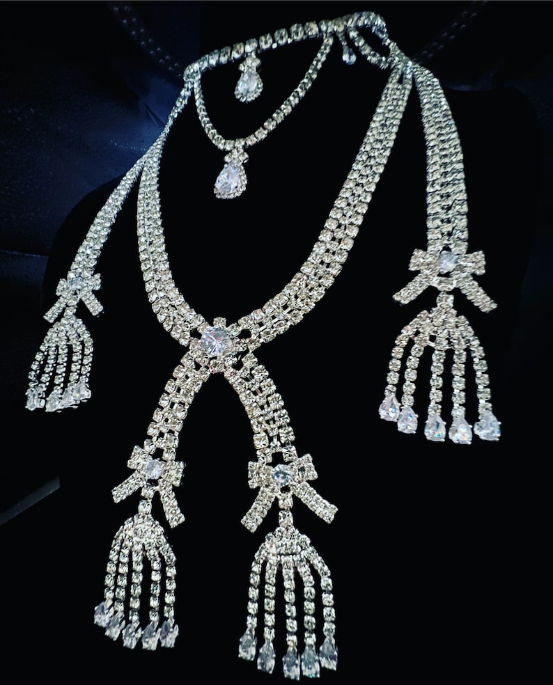May include: A silver rhinestone necklace set with three strands and three dangling bows. Each bow has a tassel of rhinestones.