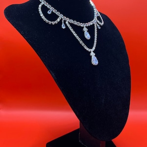 May include: A silver rhinestone choker necklace with a teardrop pendant. The necklace is displayed on a black velvet stand against a red background.