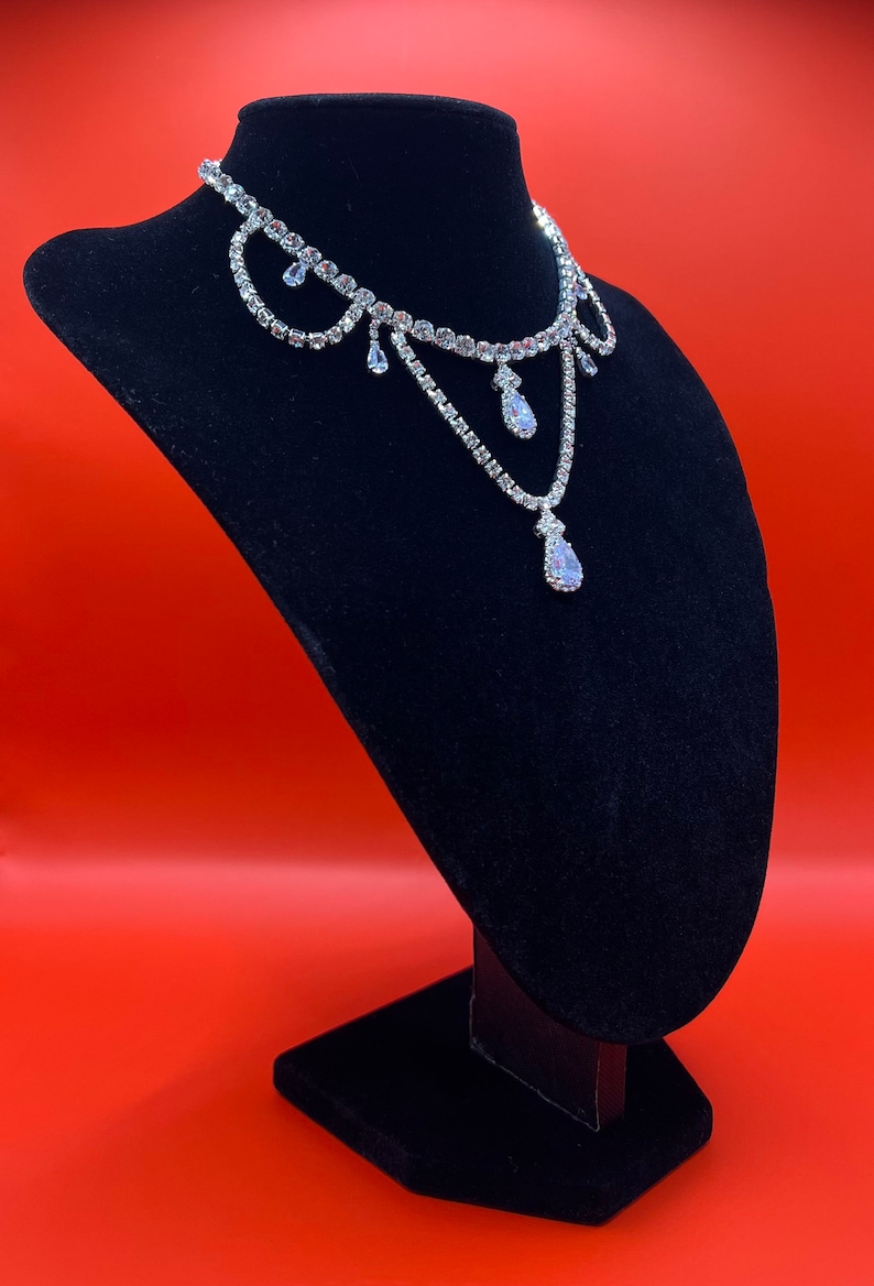 May include: A silver rhinestone choker necklace with a teardrop pendant. The necklace is displayed on a black velvet stand against a red background.