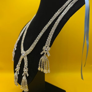 May include: A silver rhinestone necklace with two long strands and three dangling tassels. The necklace is displayed on a black velvet stand with a light blue ribbon.