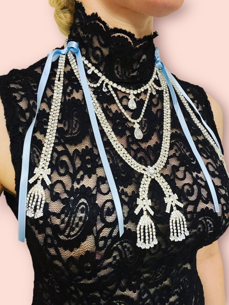May include: A black lace top with a layered rhinestone necklace and light blue ribbon ties. The necklace has two dangling rhinestone tassels.