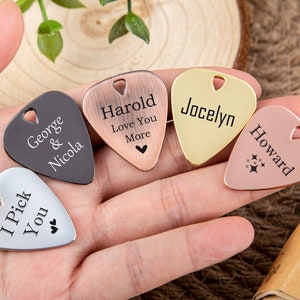 May include: Five guitar picks with personalized engravings. The picks are in different colors: black, copper, gold, silver, and rose gold. The engravings include names, messages, and a star design.