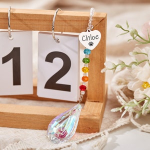 Puede incluir: Un atrapasol de cristal arco&iacute;ris con un dije de coraz&oacute;n plateado que dice "Chloe" y una huella de pata. El atrapasol est&aacute; colgado de un calendario de madera con los n&uacute;meros 11 y 12 visibles.