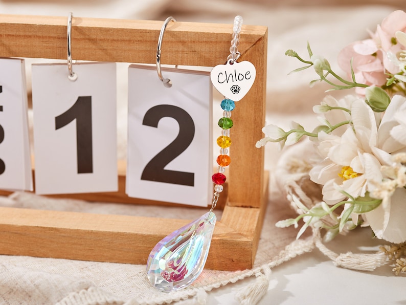 Puede incluir: Un atrapasol de cristal arco&iacute;ris con un dije de coraz&oacute;n plateado que dice "Chloe" y una huella de pata. El atrapasol est&aacute; colgado de un calendario de madera con los n&uacute;meros 11 y 12 visibles.