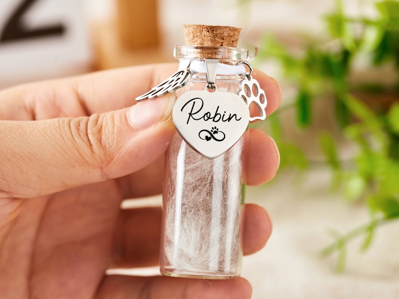 K&ouml;nnte beinhalten: Ein kleines Glasgef&auml;&szlig; mit einem Korkverschluss, gef&uuml;llt mit wei&szlig;em Fell. Das Gef&auml;&szlig; hat einen silbernen Herzanh&auml;nger mit dem Namen "Robin" und einem Unendlichkeitszeichen mit einem Pfotenabdruck. Der Anh&auml;nger ist an einem wei&szlig;en Band befestigt.