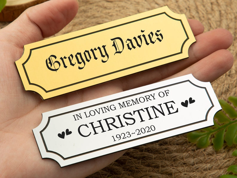 May include: Two rectangular memorial plaques. One is gold with the name "Gregory Davies" in black gothic font. The other is silver with the inscription "IN LOVING MEMORY OF CHRISTINE 1923-2020" and two heart symbols.