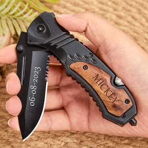 May include: A black folding knife with a wood handle. The handle is engraved with the name "Mickey" and the date "06-08-2023".