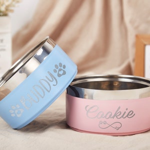 K&ouml;nnte beinhalten: Zwei personalisierte Edelstahl-Napf f&uuml;r Haustiere. Die blaue Sch&uuml;ssel sagt "Buddy" und die rosa Sch&uuml;ssel sagt "Cookie".