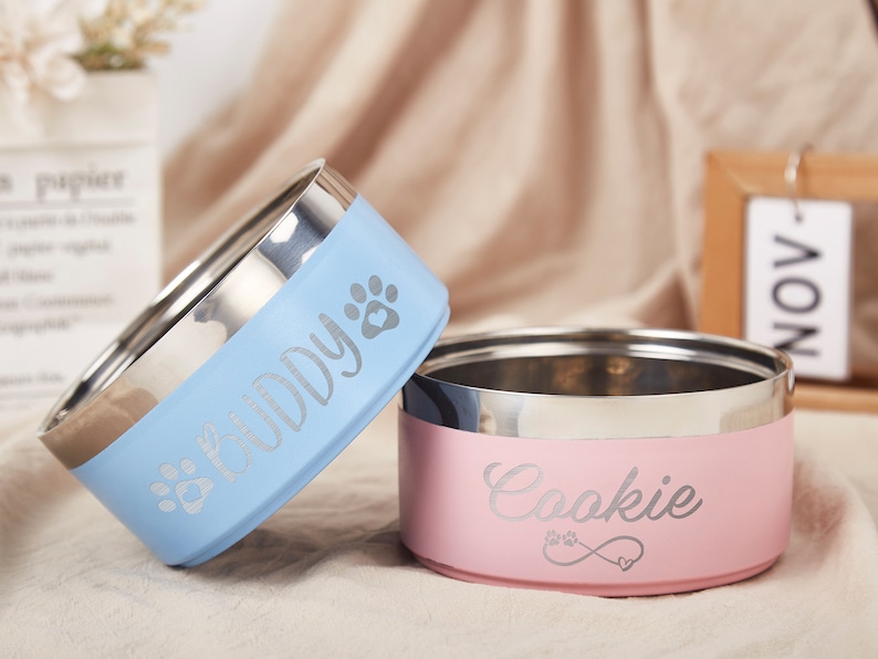 K&ouml;nnte beinhalten: Zwei personalisierte Edelstahl-Napf f&uuml;r Haustiere. Die blaue Sch&uuml;ssel sagt "Buddy" und die rosa Sch&uuml;ssel sagt "Cookie".