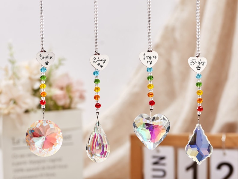 Puede incluir: Cuatro atrapasoles de cristal arco&iacute;ris con encantos personalizados en forma de coraz&oacute;n. Los encantos est&aacute;n grabados con los nombres "Sophie", "Daisy", "Jasper" y "Bailey" y una huella de pata. Cada atrapasol presenta una forma de cristal diferente.