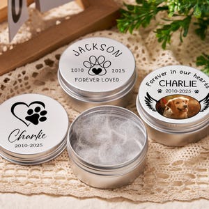 May include: Silver pet memorial tins with lids. One tin is open, filled with white fluff. The lids feature paw prints, hearts, and the names "Jackson" and "Charlie" with dates. One lid has a photo of a dog.