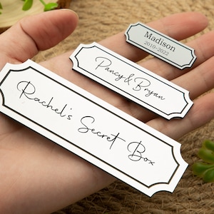 May include: Three rectangular, white and black nameplates with decorative borders. The largest plate reads "Rachel's Secret Box" in script. The other plates read "Pancy & Bryan" and "Madison 2010-2022".